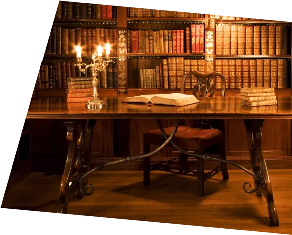 En Schreibtisch mit augeschlagenem Buch, Kerzen. Im Hintergrund Bücherregale mit alten Büchern.